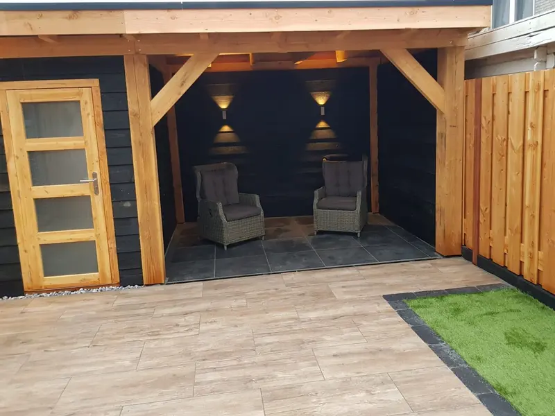 Overkapping met plat dak en een berging met houten deur met vier glasvlakken, zwarte achterwand met wandlampen, twee loungestoelen, rechts een houten schutting