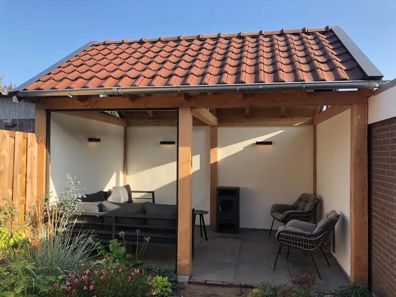 Overkapping met rood pannendak, houten staanders en schoren, lichte wanden met wandlampen, een loungeset en een houtkachel, met een border met planten ervoor