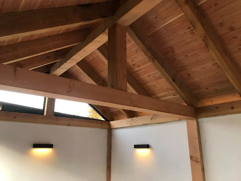 Binnenkant van de overkapping: houten spanten, schoren en dakbeschot, een glazen strook in de nok en twee brandende wandlampen