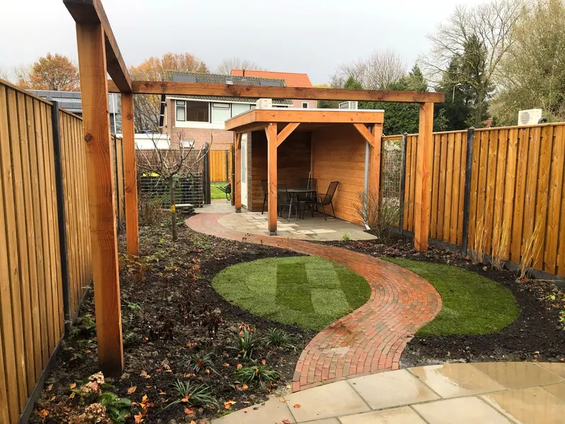 Diepe tuin met links en rechts een houten schutting, een houten pergola over een gebogen pad en achterin een overkapping met tafel en stoelen