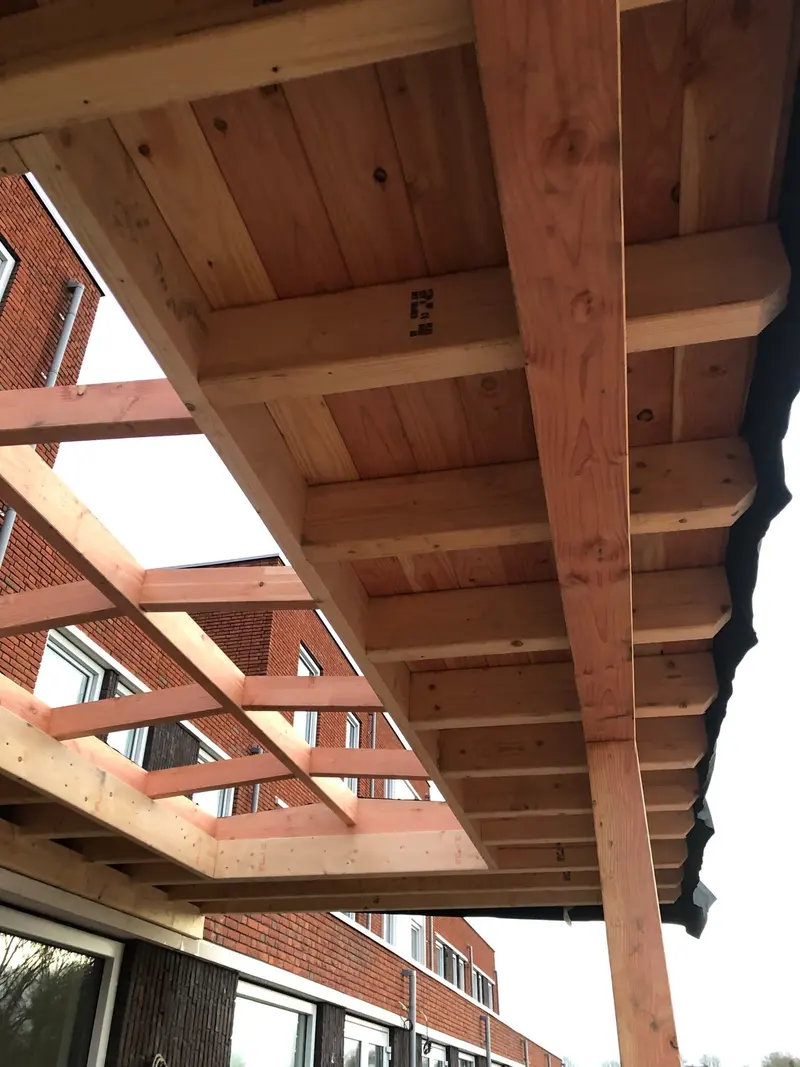 Onderaanzicht van een houten overkapping tegen een rij woningen, met balken, dakbeschot en een open raamwerk van liggers