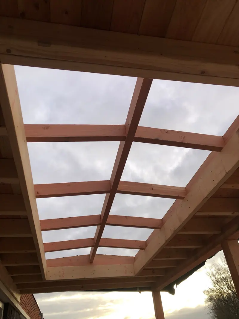 Open raamwerk van houten liggers in een overkapping, met de lucht erdoorheen