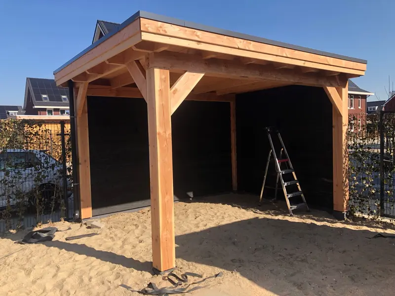 Overkapping in aanbouw met plat dak, houten staanders en schoren en twee zwarte zijwanden, op zand met een trapje eronder
