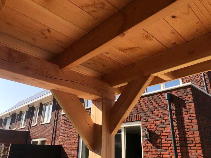 Onderaanzicht van een houten staander met twee schoren onder balklaag en dakbeschot, met bakstenen woningen op de achtergrond