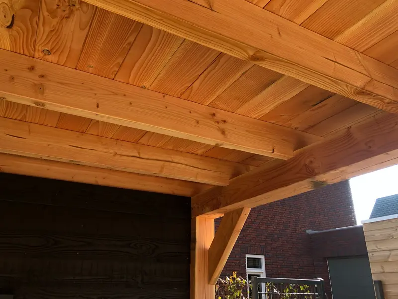 Onderkant van een houten overkapping met balklaag en dakbeschot met duidelijke nerf en noesten, boven een zwarte wand