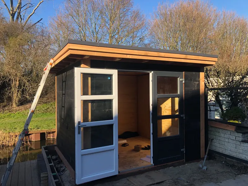 Tuinhuis met plat dak, zwarte wanden, een witte en een zwarte deur met glas en houten boeidelen, met een ladder ertegen