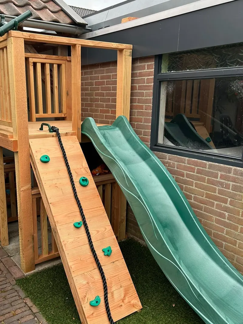 Houten speeltoestel met een platform met spijlen, een klimwand met groene grepen en een touw, en een groene glijbaan, op kunstgras naast een woning