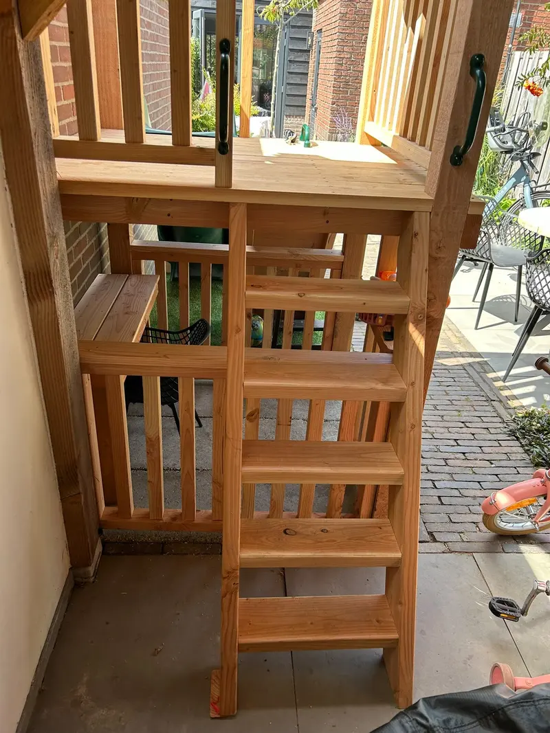 Zijkant van hetzelfde houten speeltoestel met een trap met brede treden, het platform en handgrepen